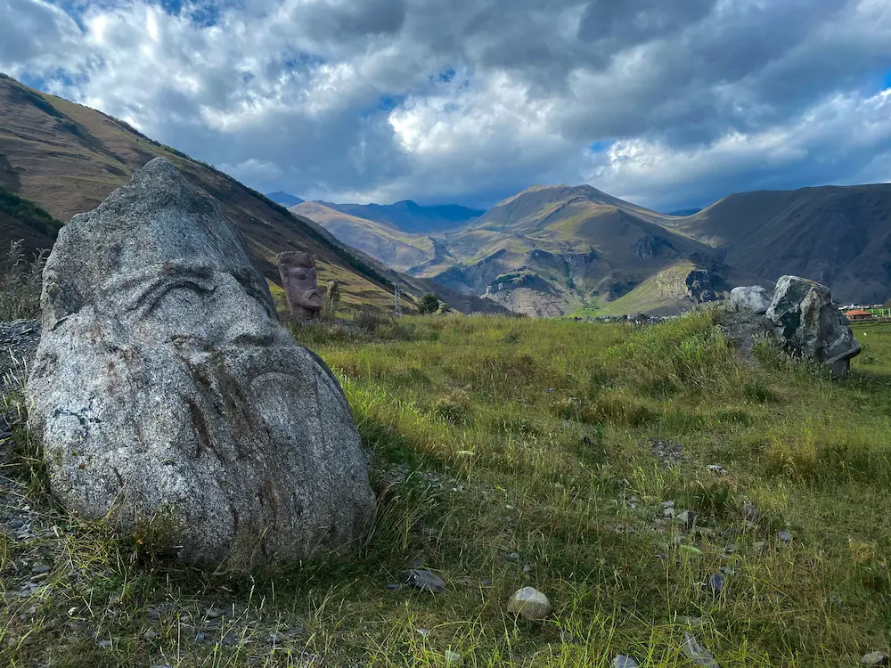 Stone sculptures with carved faces on a green hillside with the Caucasus Mountains in the background